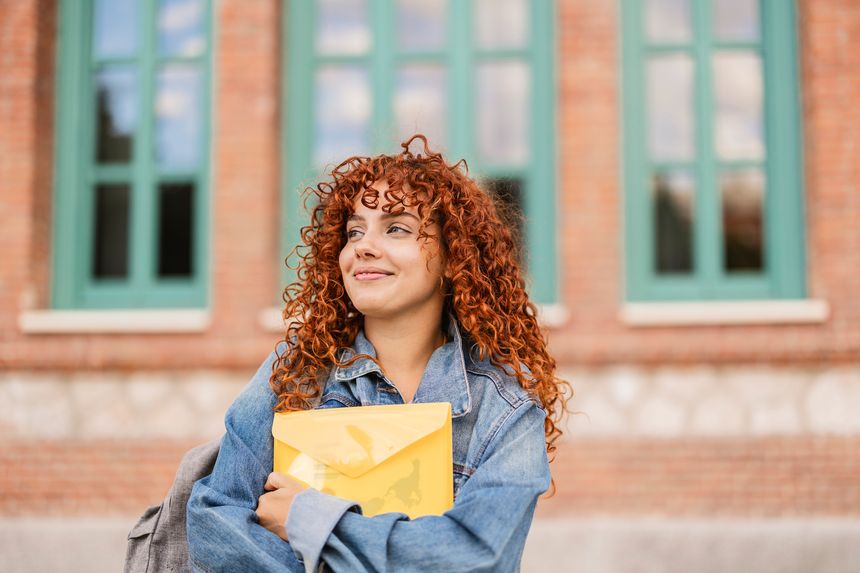 Young female student with curly red hair smiling while holding a yellow envelope containing her university acceptance letter, radiating joy and excitement on a sunny day outdoors