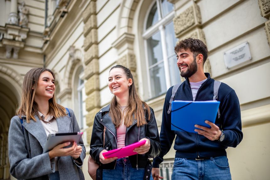 Three students stand together outdoors, holding books and literature, engaging in a lively conversation filled with laughter. Dressed casually and carrying backpacks.