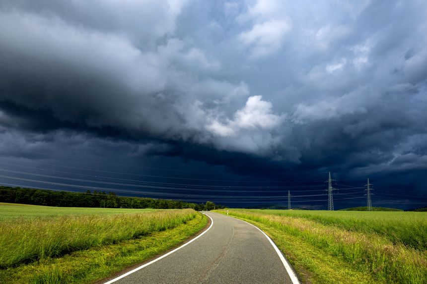 Curving rural road under dramatic storm clouds with green fields and power lines, showcasing a striking contrast of nature and infrastructure.