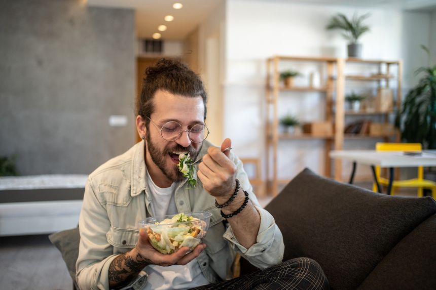 Young man with glasses and tattoos, sitting comfortably on a sofa, enjoying a fresh and vibrant salad from a plastic bowl, embracing a healthy lifestyle in his modern apartment