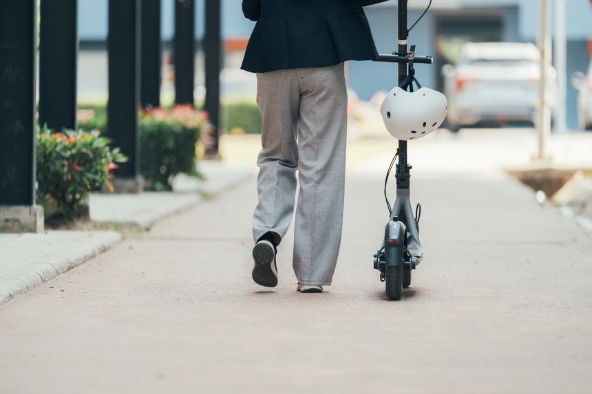 Businesswoman walking on the sidewalk while pushing an electric scooter after a long day at work, wearing a helmet for enhanced safety and promoting sustainable commuting