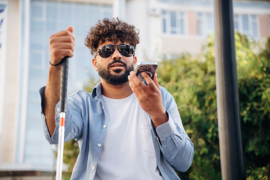 Communication, holding smartphone. Blind man with white cane and sunglasses is in the city.