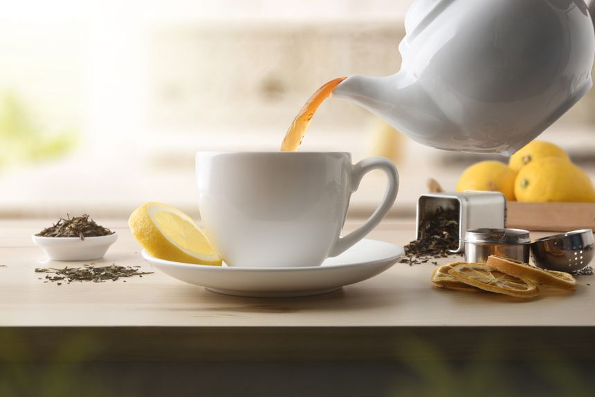 Details of serving tea from a ceramic teapot into a white ceramic cup with a slice of lemon on a wooden table with ingredients and dried tea leaves. Front view.