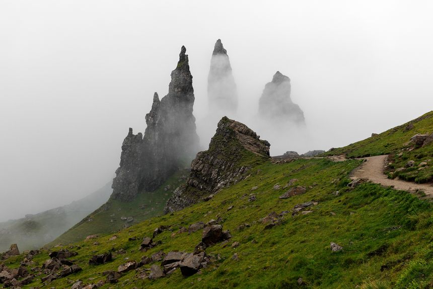 Towering black rock spires emerging from dense white fog on a steep grassy hillside in the Scottish Highlands.