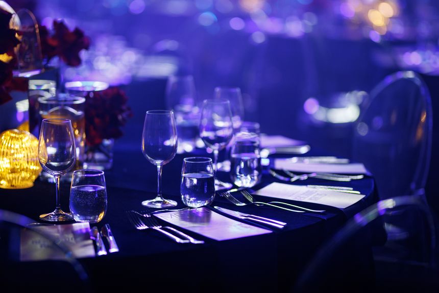 Formal dinner table with crystal glassware, polished cutlery, and menus under atmospheric blue stage lights.