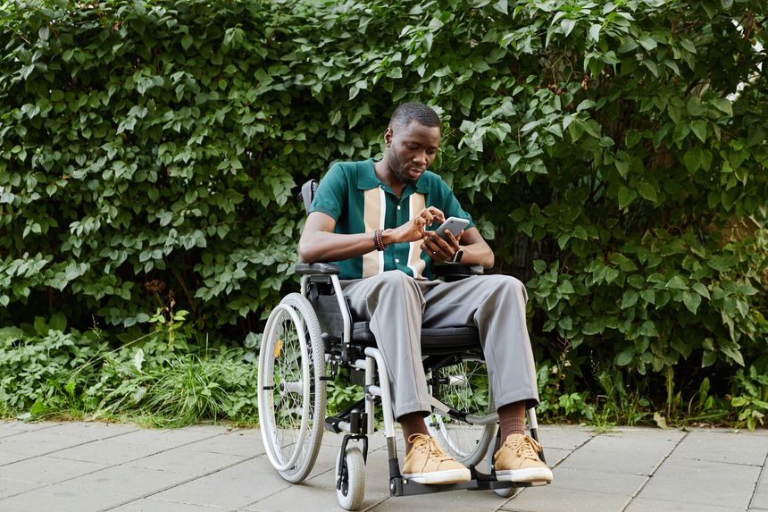 Full length portrait of adult man with disability using smartphone outdoors and zooming in photo while sitting in wheelchair outdoors against green background copy space