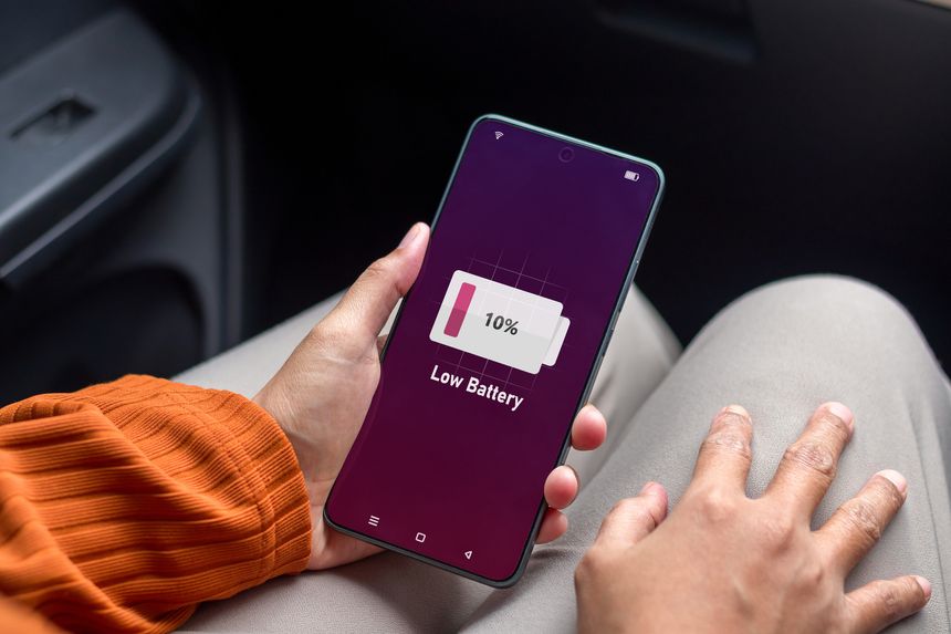Running out of battery while on the go. Woman in car holding a mobile phone with low battery symbol on screen.