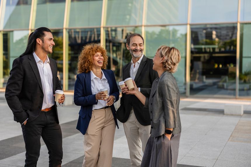 Diverse business professionals chat and laugh outdoors over coffee cups, sharing a friendly, informal break that highlights teamwork, networking and positive collaboration in the city
