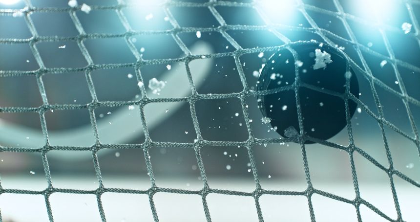 Action sports photo of a hockey puck flying into the goal net during a scoring shot, with ice particles in the air and a hockey stick visible in the background, capturing speed and intensity.