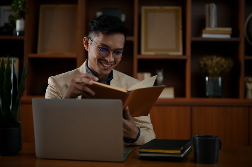 Smart and happy millennial Asian businessman reading a book at his desk in the office room.
