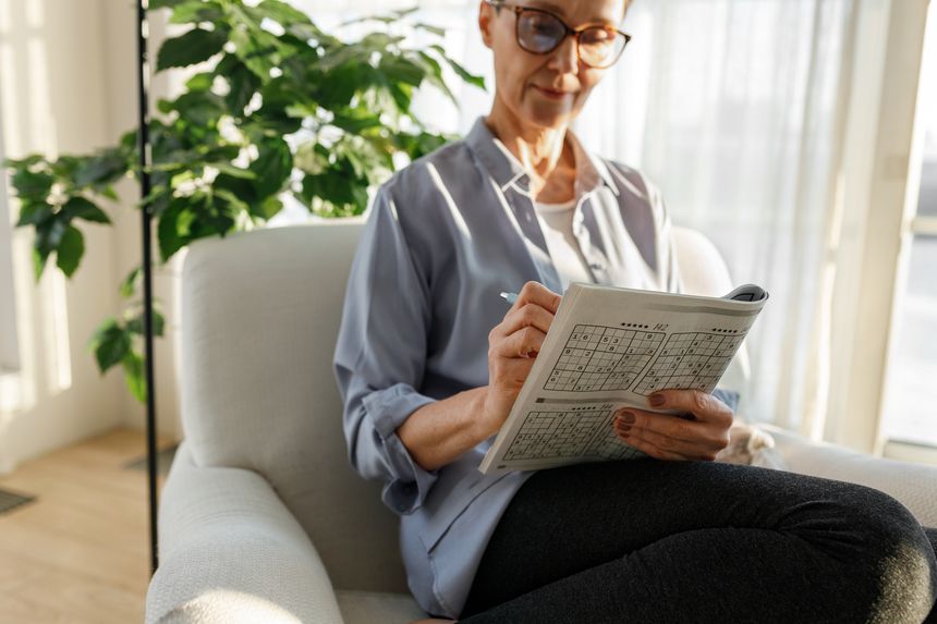 Selective focus on hands of intelligent senior lady in casual clothes and glasses holding book with puzzle games and pen, enjoying solving sudoku as preventive strategy against dementia
