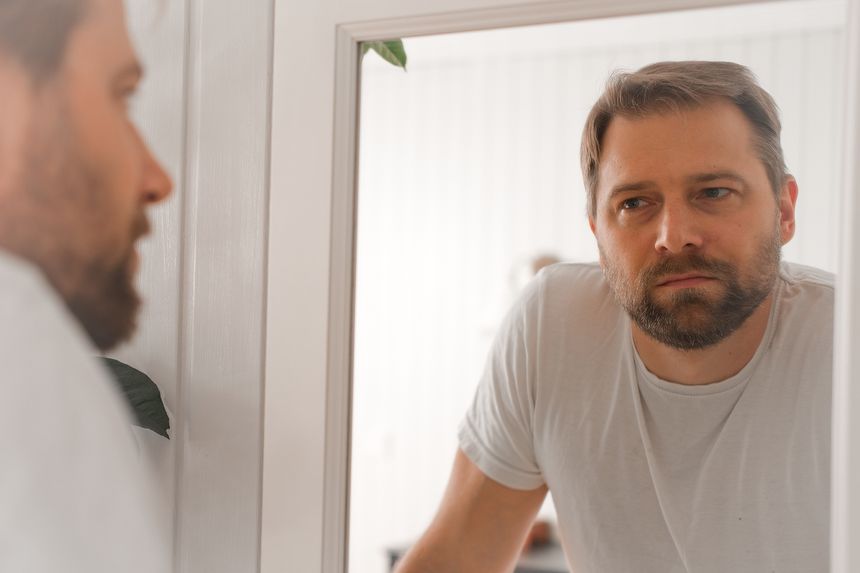 Confident handsome bearded man looking himself at the mirror. Pensive guy gazing thoughtfully. Captures introspection, grooming, hair care. Suitable for male health, lifestyle or personal care content