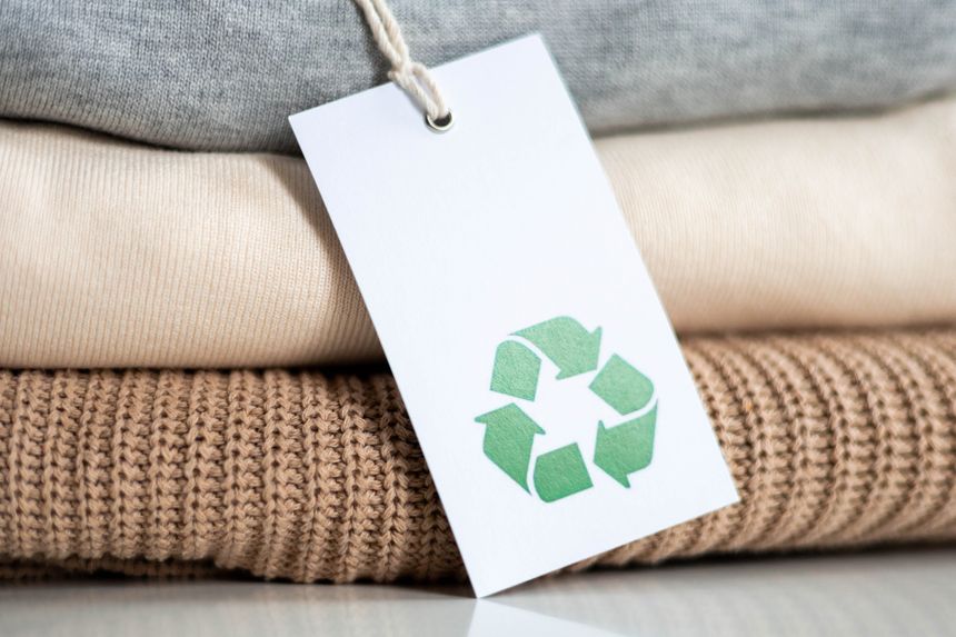 Stack of knitted clothes in neutral tones made of natural materials lies on table. Recyclable product concept. Organic cotton recycling fabric. Label with recycling icon.