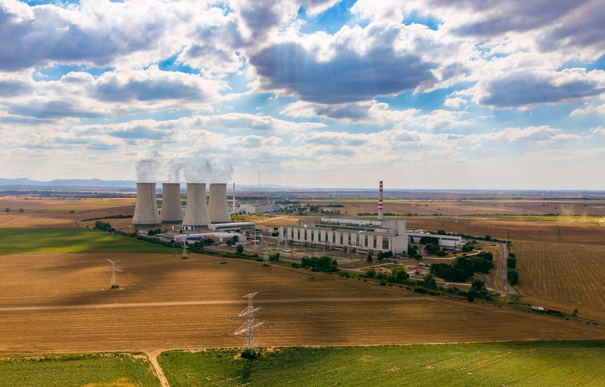 Aerial view of nuclear power plant surrounded by agricultural fields. Four concrete cooling towers emitting steam stand near main building complex under cloudy sky.