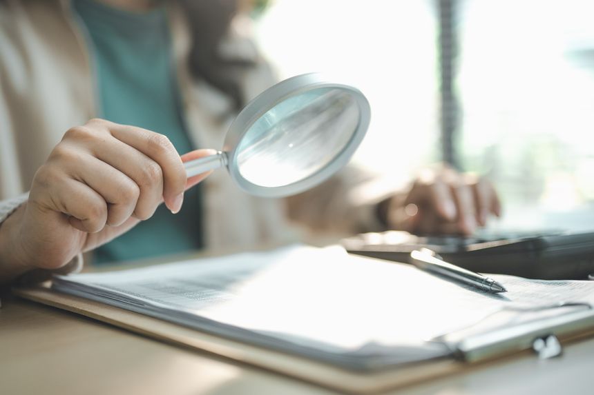 Professional reviewing business documents with magnifying glass at office desk, representing document management, audit, compliance verification, financial review and corporate inspection process.