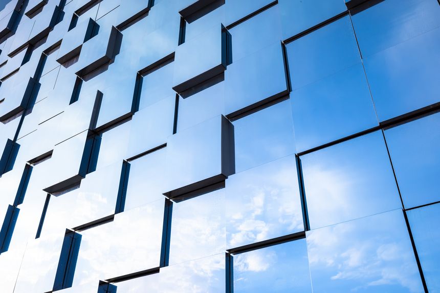 Contemporary glass facade of a commercial building featuring repeating geometric windows and reflective panels that mirror the blue sky, creating an abstract urban pattern and minimalist architecture