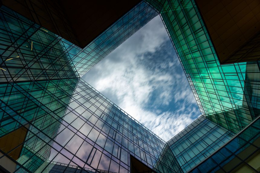 Budapest, Hungary - July 26, 2025: A striking symmetrical composition looking upward at a modern office building. The glass panels create complex reflections of moving clouds, while teal and green accents add a futuristic feel. This high-contrast architectural shot is perfect for corporate branding, technology backgrounds, and real estate marketing, offering a clean central copy space for text.