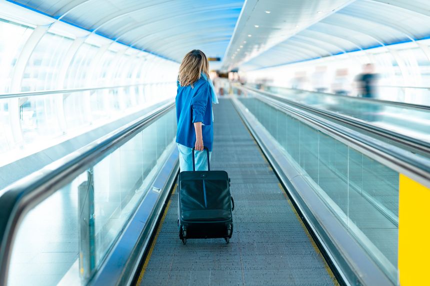 A person with shoulder-length hair walks through a spacious airport walkway, pulling a suitcase behind them, surrounded by travelers in a bright and airy terminal.