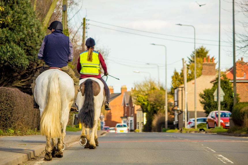 March 25, 2018 : Kermington, England, United Kingdom.  Two female horse riders, seen from the back, on their horses.  The horses walking at the side of a village road, tucked into the side where the kerb of the road it.