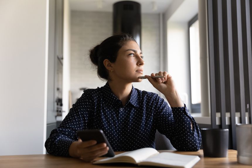 Pensive young Indian woman write in notebook use cellphone look in distance pondering thinking. Thoughtful millennial female plan decide make notes working on smartphone at home office.