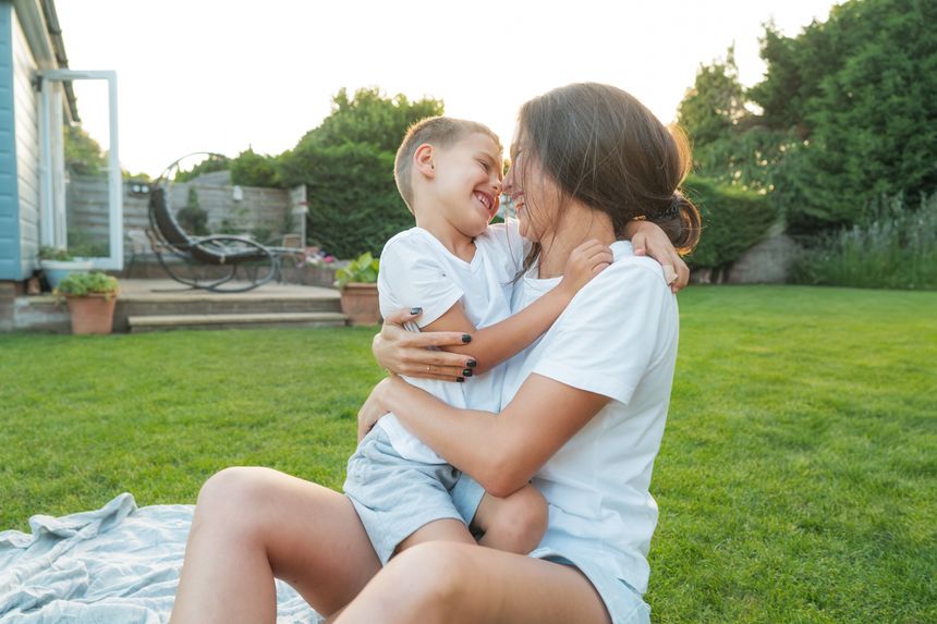Cheerful cute preschooler boy kid having fun with his mum, embracing, laughing on the green grass. Young mom and kid hugging with love, affection, tenderness on the backyard lawn background on sunset.