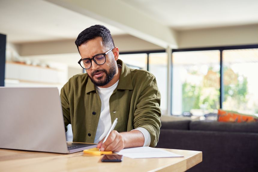 Young business man working on laptop and writing on sticky notes at home. Happy indian businessman working on laptop while writing notes in sticky pad at home with copy space. Focused multiethnic man working from home analyzing financial data and doing research.