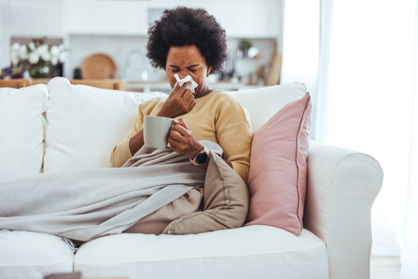 Shot of a mature woman blowing her nose with a tissue at home. Woman sneezing in a tissue in the living room. Worried African American woman sitting at home while being sick