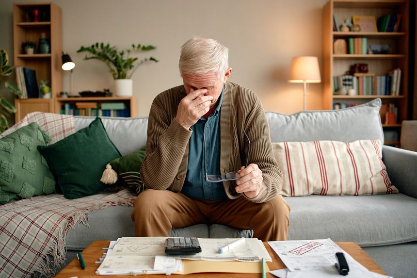 Senior man sitting on sofa analyzing invoices and bills, holding eyeglasses in one hand and rubbing forehead with other hand, paperwork and calculator on table