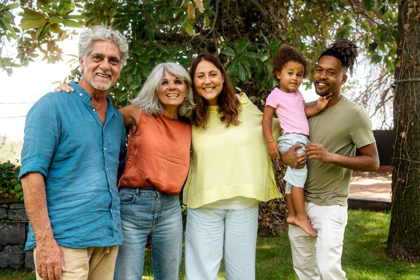 Diverse, multigenerational family portrait - moment of affection - Grandparents beam with joy next to their daughter and son-in-law, who holds a toddler. Child touching her father's face.