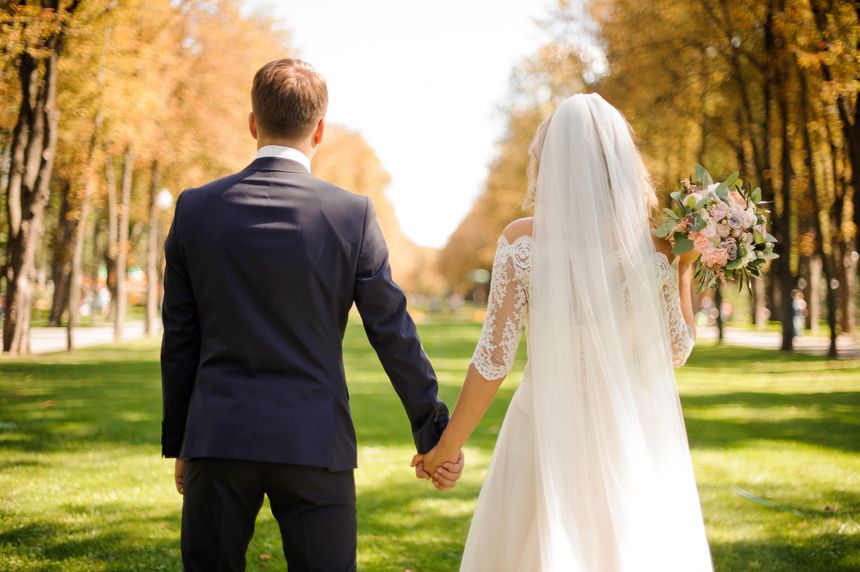 Back view of bride and bridegroom holding hands on the park lawn against the background of yellow trees