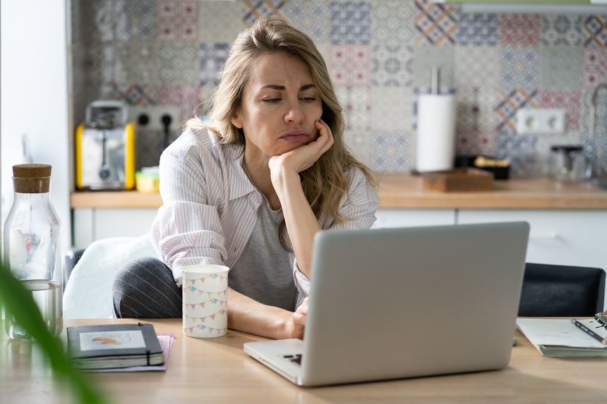 Unhappy woman of middle age search for new job unemployed during covid-19 quarantine. Female look at laptop screen with vacancies sitting unpleased on home kitchen in pajama. Unemployment concept
