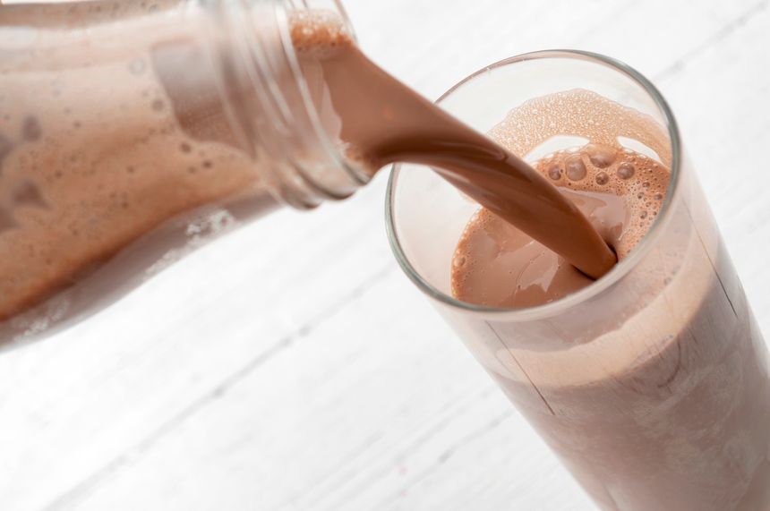 Calcium and protein rich beverage, nutritious milkshake and refreshing treat concept with photograph of bottle pouring chocolate milk into a tall glass isolated on rustic white wood background