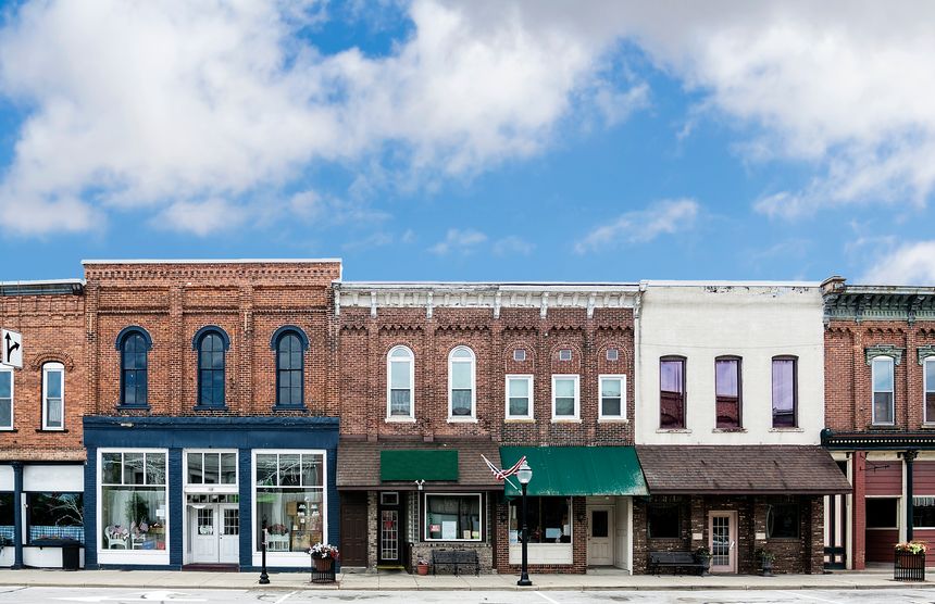 A photo of a typical small town main street in the United States of America. Features old brick buildings with specialty shops and restaurants. Decorated with spring flowers and American flags.