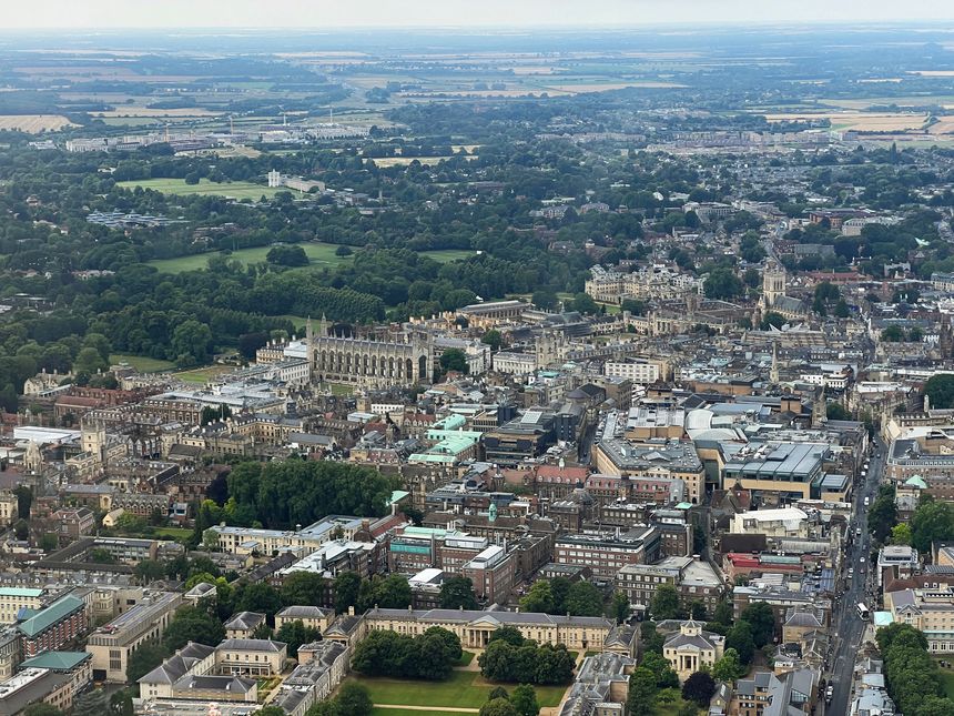 Aerial view of Cambridge, England, featuring historic university buildings, college courtyards, and a dense urban layout surrounded by green spaces and countryside.