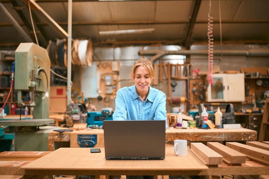 Female Carpenter Working In Woodwork Workshop Using Laptop With Cup Of Coffee
