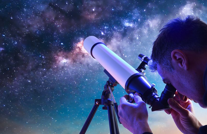 Detail of man outdoors looking through the eyepiece of a telescope focusing on a blue sky full of stars.