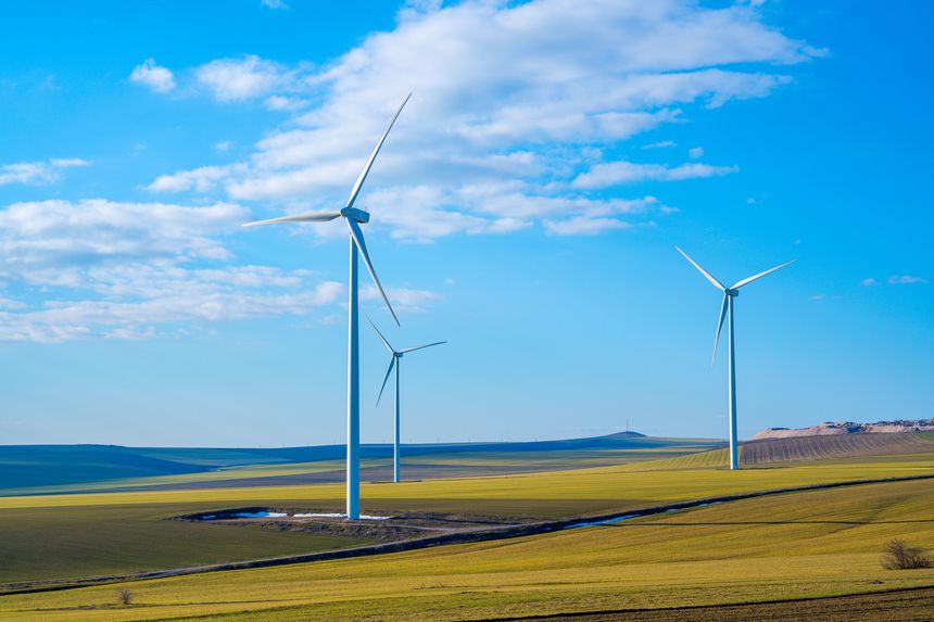 Several wind turbines are placed in an open field. The landscape shows green grass and small hills. Bright blue sky with scattered clouds surrounds the scene.