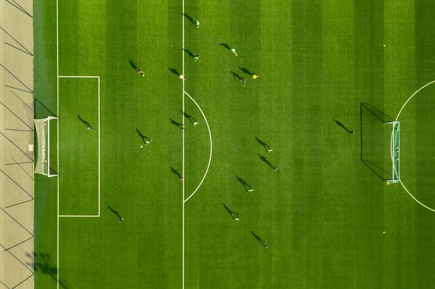 Aerial view players playing match stadium below large green football field with white lined lines. Playing football at stadium. Professional soccer players are located on playing field. Sport
