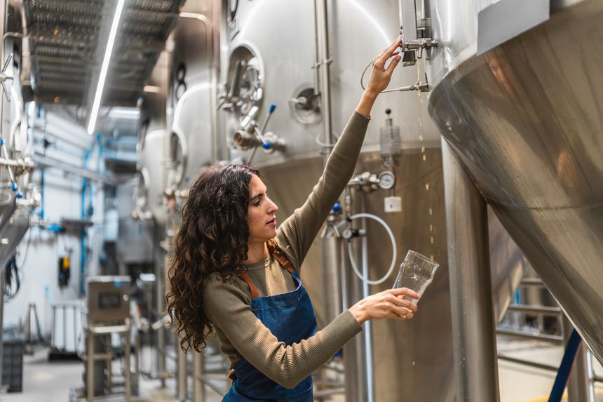 Female worker in denim apron checking quality control of craft beer from a fermentation tank in a modern industry