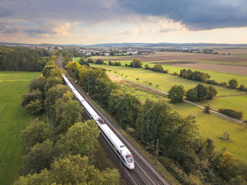 A high-speed train travels through lush green countryside with a village in the background under a dramatic sky.