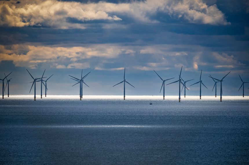 Offshore wind farm set against a dramatic sky with dense clouds. A narrow strip of sunlight illuminates the ocean surface in front of the wind turbines, creating a striking contrast. The turbines are evenly spaced, with large blades harnessing wind energy. The deep blue sea is calm, and the atmosphere suggests a peaceful, yet dynamic environment. An isolated small boat appears near the turbines, highlighting the scale of the scene.