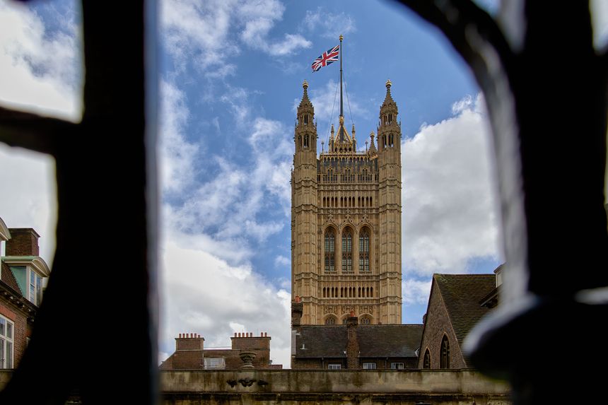 View of the Victoria Tower, part of the UK Parliament, framed through a window with a Union Jack flag flying, under a partly cloudy sky.