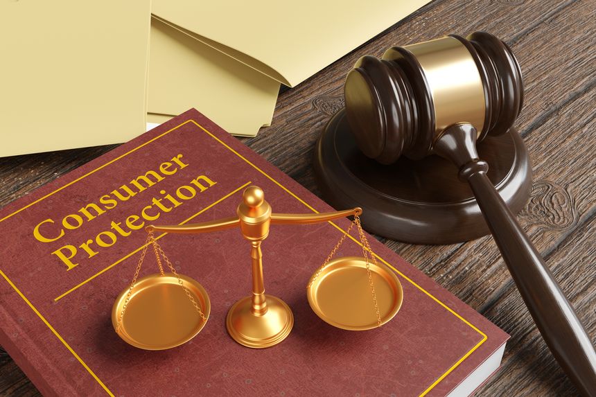 Golden scales of justice on a brown leather bound book engraved with the title Consumer Protection, together with a gavel and yellow office folders on a wooden table