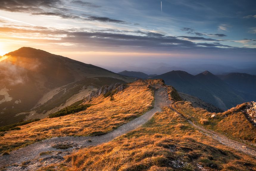 A mountain path stretches across a ridge at sunset. The sky displays a mix of clouds and sunlight. Distant mountain peaks are visible in the background.