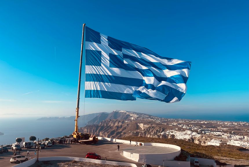 The Greek flag flies high against a clear blue sky, overlooking a stunning coastal landscape, symbolizing the nation's heritage and beauty.