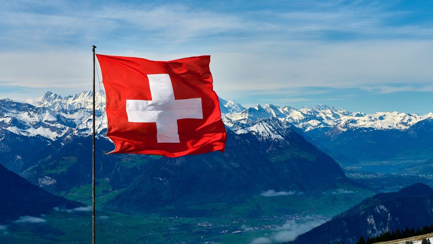 High above the valleys, the Swiss flag flutters proudly. Majestic mountains stretch across the horizon while the bright blue sky enhances the stunning scenery, showcasing the beauty of Switzerland.