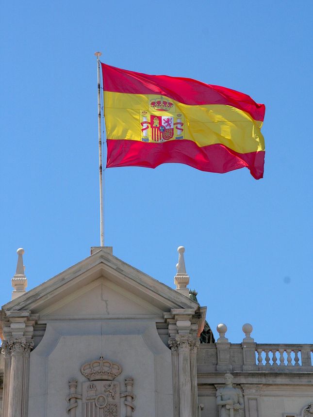 A bold red-and-yellow Spanish flag ripples in the breeze above a stately building, its colors vivid against the sky. Sunlight catches the coat of arms at the flag’s center, emphasizing its royal gold and crimson details. Below, the structure’s stone façade — with classic balconies and wrought-iron railings — speaks to Spain’s architectural heritage, framing the flag as a proud national emblem.