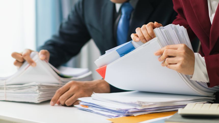 Two businesswomen are working together to search through important documents from a pile of documents on a table to verify the authenticity of the documents that were previously signed in a contract.