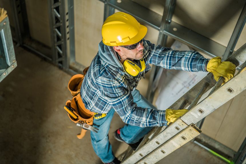 Top View Photo of Professional Contractor in Yellow Hard Hat Climbing the Ladder at the Construction Site. Building Tools and Equipment in Use.