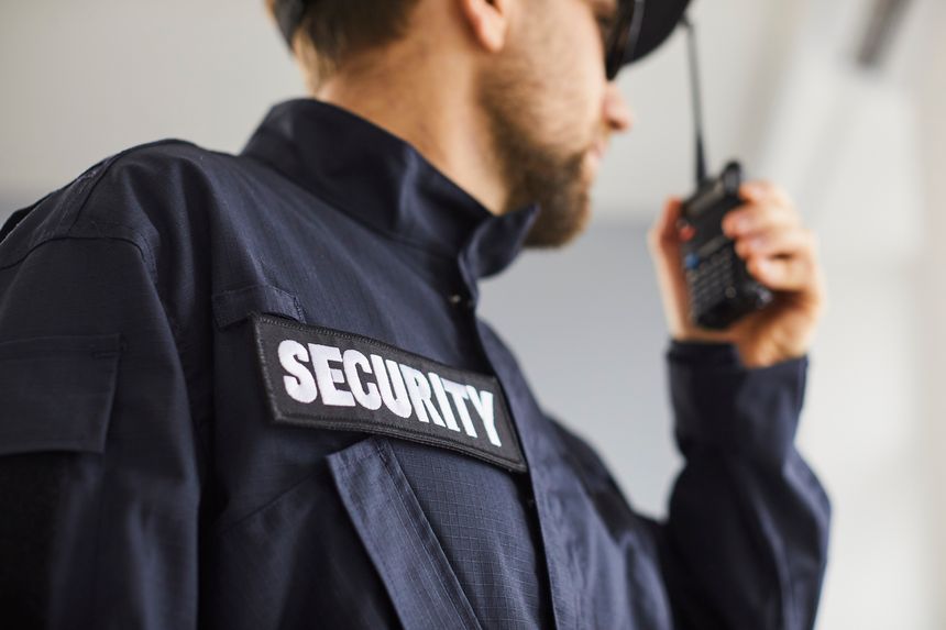 Close up cropped portrait of confident male security guard standing indoors. Bearded guardian man in blue uniform talking by the walkie talkie and looking to the side. Selective focus.